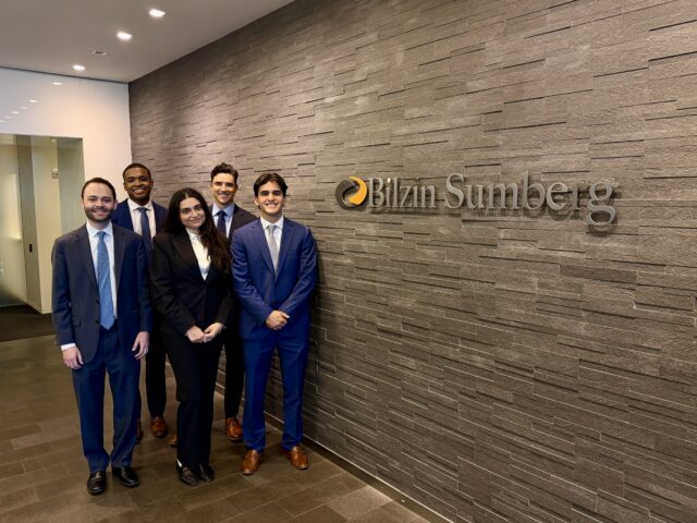 Five summer associates standing in the Bilzin Sumberg office lobby in business attire, posing in front of the firm’s wall logo. The group is smiling and dressed in suits, with a modern stone accent wall and office hallway visible in the background.